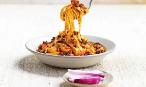 Un piatto invitante di tagliatelle fatte in casa con ragù ricco, sollevate da una forchetta. Accanto, spicchi di cipolla rossa fresca.