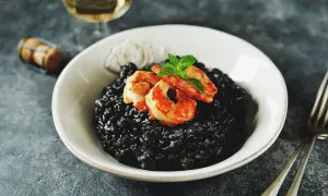 Un invitante risotto al nero di seppia, guarnito con gamberi succulenti e menta fresca, pronto per essere gustato con un bicchiere di vino bianco.