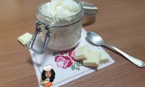 Un vasetto pieno di namelaka al cioccolato bianco, con pezzi di cioccolato e un cucchiaino.
