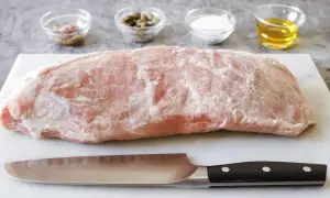 Un pezzo di magatello di vitello crudo, pronto per essere cucinato, con coltello e ciotoline di condimenti sullo sfondo.