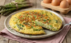 Frittata di asparagi dorata e soffice, guarnita con prezzemolo fresco. Sullo sfondo, un mazzo di asparagi e uova.