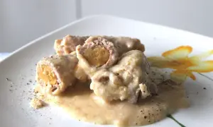 Involtini di carne succulenti in salsa cremosa, cosparsi di pepe nero su un piatto bianco decorato con un fiore giallo.