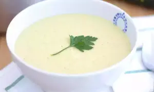 Una cremosa vellutata di zucchine, guarnita con un rametto di prezzemolo, servita in una ciotola bianca con un bordo decorato.