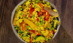 Un invitante riso al curry con piselli, peperoni rossi e gialli, zucchine e cipolla, servito in una ciotola su un tavolo di legno.