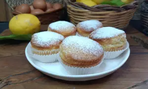 Cinque muffin allo yogurt spolverati di zucchero a velo, pronti per essere gustati. Sullo sfondo, limoni e uova fresche.
