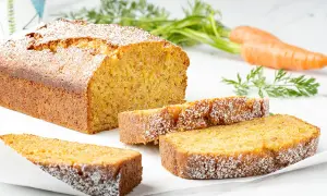 Torta carote e mandorle soffice e dorata, cosparsa di zucchero a velo. Due fette sono servite accanto al pane intero, con carote fresche sullo sfondo.