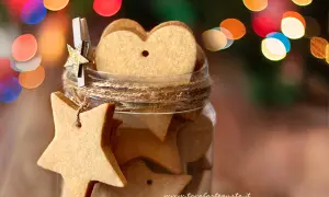 Biscotti alla cannella a forma di stella e cuore in un barattolo, decorati con luci natalizie sfocate.