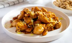Un invitante piatto di pollo alle mandorle, lucido e dorato, guarnito con erba cipollina. Sullo sfondo, una ciotola di mandorle intere.