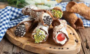 Cannoli siciliani ricetta: deliziosi dolci con crema, gocce di cioccolato, pistacchi e ciliegina candita su tagliere di legno.