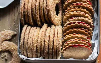 Biscotti grano saraceno ripieni di marmellata, impilati in una scatola di latta. Alcuni biscotti a forma di ciambella e a mezzaluna sono sparsi sul tavolo.