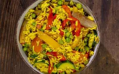 Un invitante riso al curry con piselli, peperoni rossi e gialli, zucchine e cipolla, servito in una ciotola su un tavolo di legno.