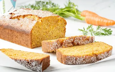 Torta carote e mandorle soffice e dorata, cosparsa di zucchero a velo. Due fette sono servite accanto al pane intero, con carote fresche sullo sfondo.