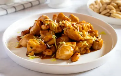 Un invitante piatto di pollo alle mandorle, lucido e dorato, guarnito con erba cipollina. Sullo sfondo, una ciotola di mandorle intere.
