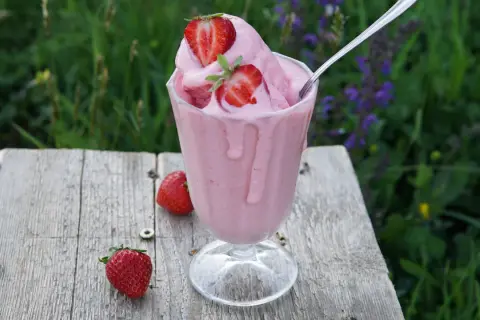Un cremoso gelato alla fragola, guarnito con fragole fresche, su un tavolo di legno rustico, con un prato fiorito sullo sfondo.