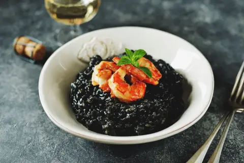 Un invitante risotto al nero di seppia, guarnito con gamberi succulenti e menta fresca, pronto per essere gustato con un bicchiere di vino bianco.