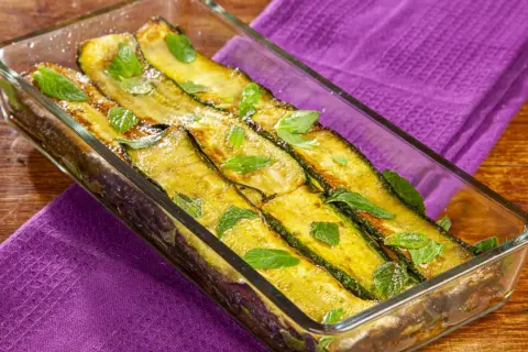 Zucchine alla scapece dorate e profumate, guarnite con foglie di menta fresca in una pirofila di vetro su un tavolo di legno.