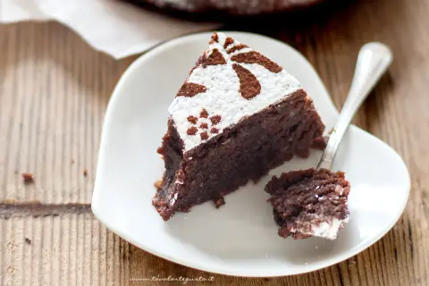 Una fetta di torta caprese ricetta facile, cosparsa di zucchero a velo e cacao, su un piatto bianco a forma di cuore.