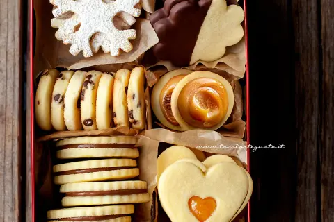 Scatola piena di biscotti al burro: a fiocco di neve, con gocce di cioccolato, ripieni di caramello, a forma di cuore e ricoperti di cioccolato.