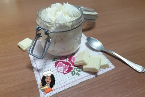 Un vasetto pieno di namelaka al cioccolato bianco, con pezzi di cioccolato e un cucchiaino.