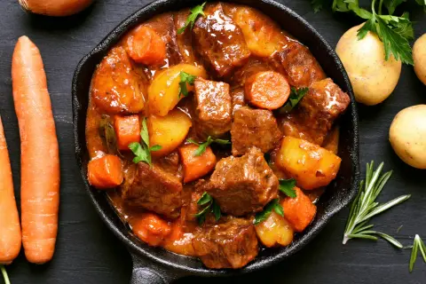 Un saporito spezzatino di maiale in padella con patate e carote, guarnito con prezzemolo fresco.