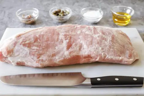 Un pezzo di magatello di vitello crudo, pronto per essere cucinato, con coltello e ciotoline di condimenti sullo sfondo.