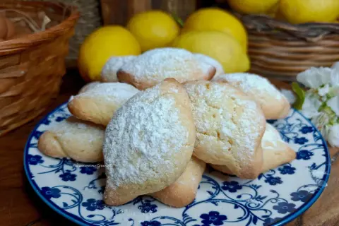Biscotti con farina di mandorle, spolverati di zucchero a velo, su un piatto decorato. Sullo sfondo limoni e un cestino.