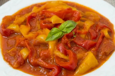 Peperonata ricetta della nonna: peperoni rossi e gialli cotti lentamente in un sugo saporito, guarniti con una foglia di basilico fresco.
