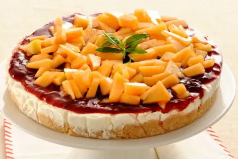 Deliziosa cheesecake al melone, con topping di frutta fresca e salsa rossa, su un piatto bianco.