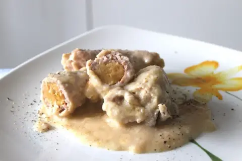 Involtini di carne succulenti in salsa cremosa, cosparsi di pepe nero su un piatto bianco decorato con un fiore giallo.