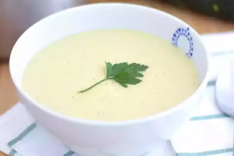 Una cremosa vellutata di zucchine, guarnita con un rametto di prezzemolo, servita in una ciotola bianca con un bordo decorato.
