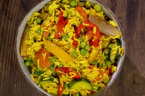Un invitante riso al curry con piselli, peperoni rossi e gialli, zucchine e cipolla, servito in una ciotola su un tavolo di legno.
