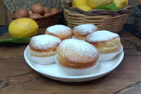 Cinque muffin allo yogurt spolverati di zucchero a velo, pronti per essere gustati. Sullo sfondo, limoni e uova fresche.