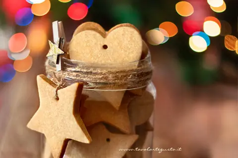 Biscotti alla cannella a forma di stella e cuore in un barattolo, decorati con luci natalizie sfocate.
