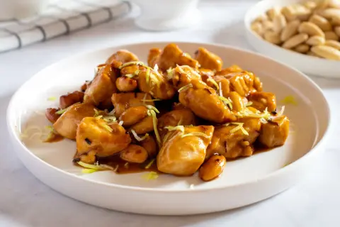 Un invitante piatto di pollo alle mandorle, lucido e dorato, guarnito con erba cipollina. Sullo sfondo, una ciotola di mandorle intere.