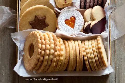 Scatola piena di deliziosi dolci senza glutine: biscotti rotondi con stella al cioccolato, a forma di cuore con zucchero a velo, ricoperti di cioccolato e impilati in file ordinate.