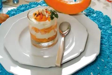 Delizioso semifreddo al melone a strati, guarnito con frutta fresca e mandorle, servito in un bicchiere su un piatto bianco.