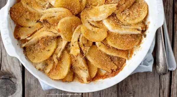 Finoçchi gratinati al forno, dorati e croccanti, in una pirofila bianca su un tavolo di legno.