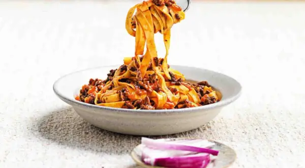 Un piatto invitante di tagliatelle fatte in casa con ragù ricco, sollevate da una forchetta. Accanto, spicchi di cipolla rossa fresca.