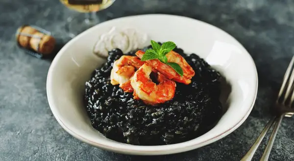 Un invitante risotto al nero di seppia, guarnito con gamberi succulenti e menta fresca, pronto per essere gustato con un bicchiere di vino bianco.