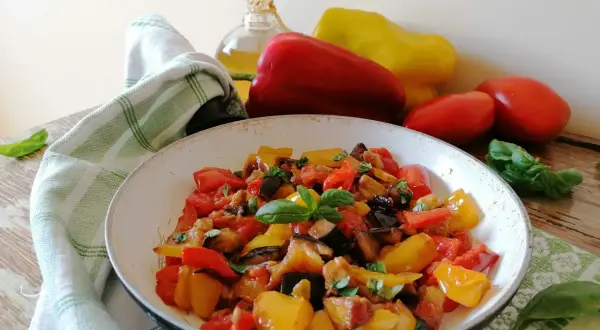 Un piatto colorato di peperonata con melanzane, pomodori e peperoni, guarnito con basilico fresco. La ricetta perfetta per un contorno estivo.