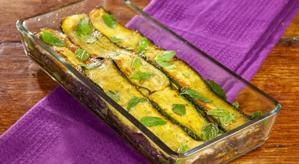 Zucchine alla scapece dorate e profumate, guarnite con foglie di menta fresca in una pirofila di vetro su un tavolo di legno.