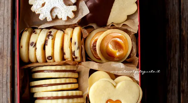Scatola piena di biscotti al burro: a fiocco di neve, con gocce di cioccolato, ripieni di caramello, a forma di cuore e ricoperti di cioccolato.