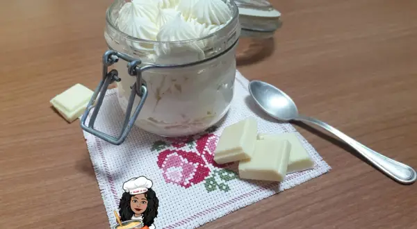 Un vasetto pieno di namelaka al cioccolato bianco, con pezzi di cioccolato e un cucchiaino.
