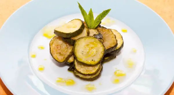 Fette di zucchine alla scapece, condite con olio e guarnite con menta fresca, su un piatto bianco.