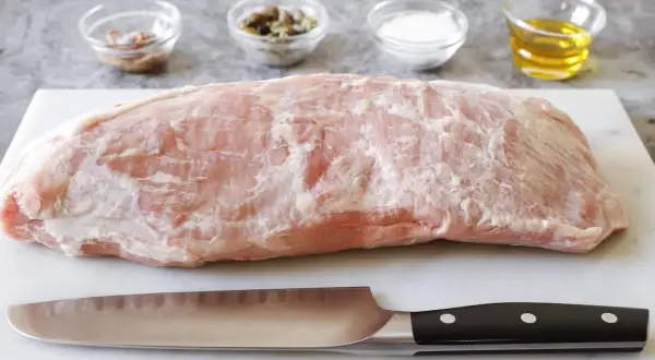 Un pezzo di magatello di vitello crudo, pronto per essere cucinato, con coltello e ciotoline di condimenti sullo sfondo.