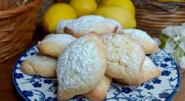 Biscotti con farina di mandorle, spolverati di zucchero a velo, su un piatto decorato. Sullo sfondo limoni e un cestino.