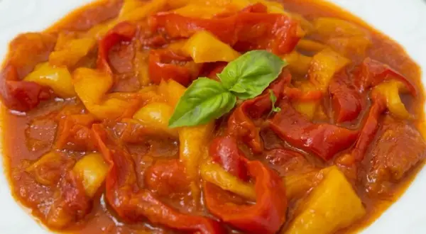 Peperonata ricetta della nonna: peperoni rossi e gialli cotti lentamente in un sugo saporito, guarniti con una foglia di basilico fresco.