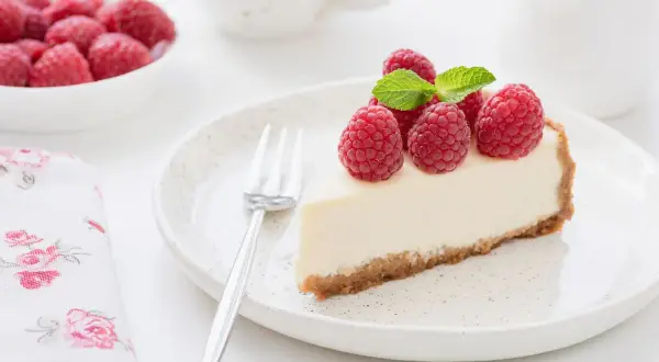 Una fetta di torta fredda allo yogurt, guarnita con lamponi freschi e menta, su un piatto bianco.
