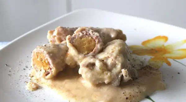 Involtini di carne succulenti in salsa cremosa, cosparsi di pepe nero su un piatto bianco decorato con un fiore giallo.