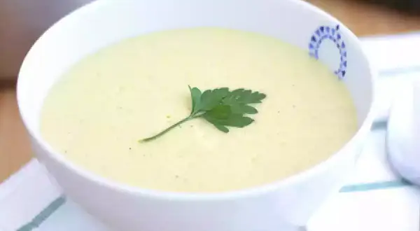 Una cremosa vellutata di zucchine, guarnita con un rametto di prezzemolo, servita in una ciotola bianca con un bordo decorato.